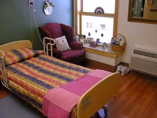 A small, cozy bedroom in a care facility featuring a single bed with a colorful striped bedspread and a pink blanket at the foot. Next to the bed is a maroon armchair with a pillow, a window with a wide sill displaying framed photos and decorative items, a basket, and a heating/cooling unit below the window. The floor is wooden, and the walls are painted green.