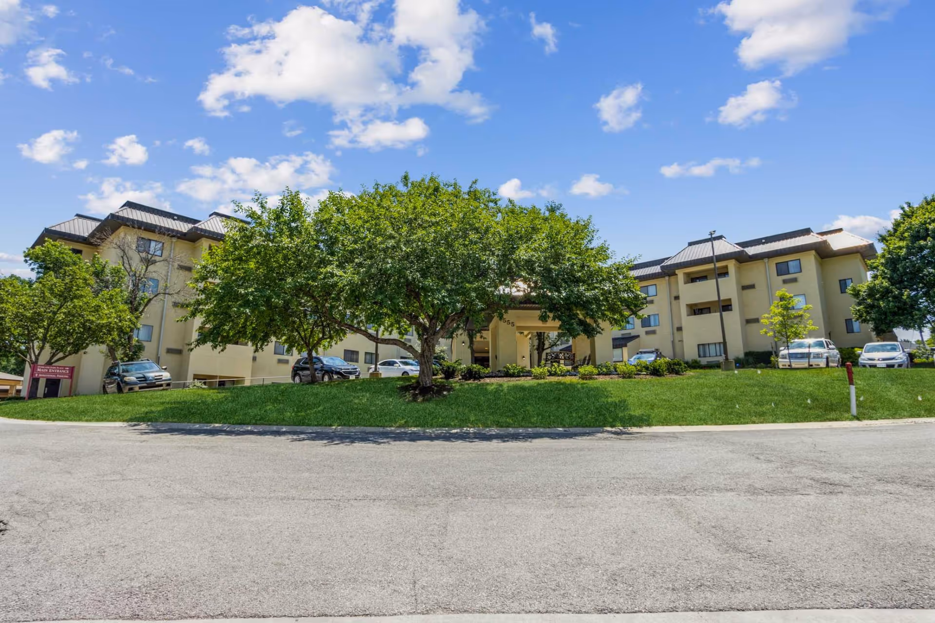 Front exterior of a multi-story senior living building with trees, parked cars, and a grassy lawn under a blue sky.