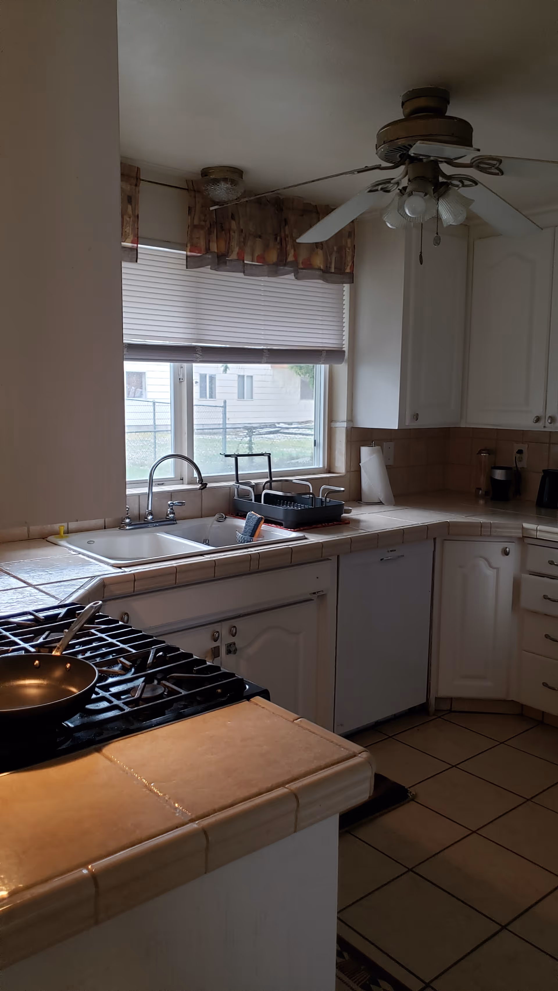 A kitchen with white cabinets, a tiled countertop, a gas stove with a frying pan, a double sink under a window with blinds and a valance, a ceiling fan with lights, and various kitchen items on the counter including a dish rack and paper towel holder.