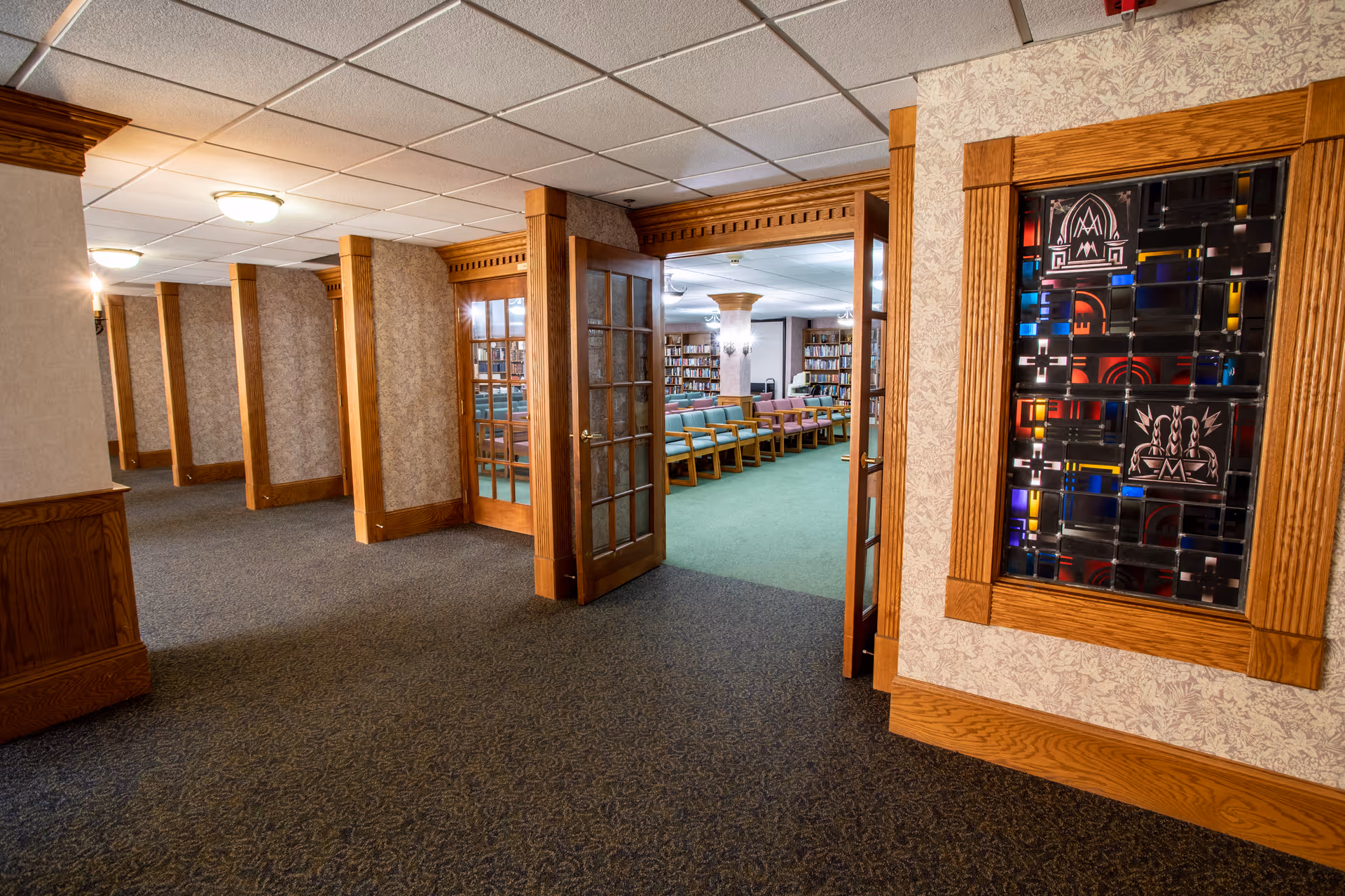 Interior view of a senior living facility hallway with carpeted floors and wood-trimmed walls. Double glass doors open into a room with rows of chairs and bookshelves along the walls. A decorative stained glass panel is mounted on the right wall near the entrance to the room.