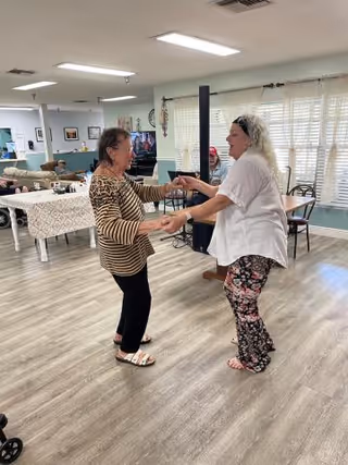 Two women holding hands and dancing together in a spacious room with wooden flooring, tables, chairs, and large windows with blinds in the background.