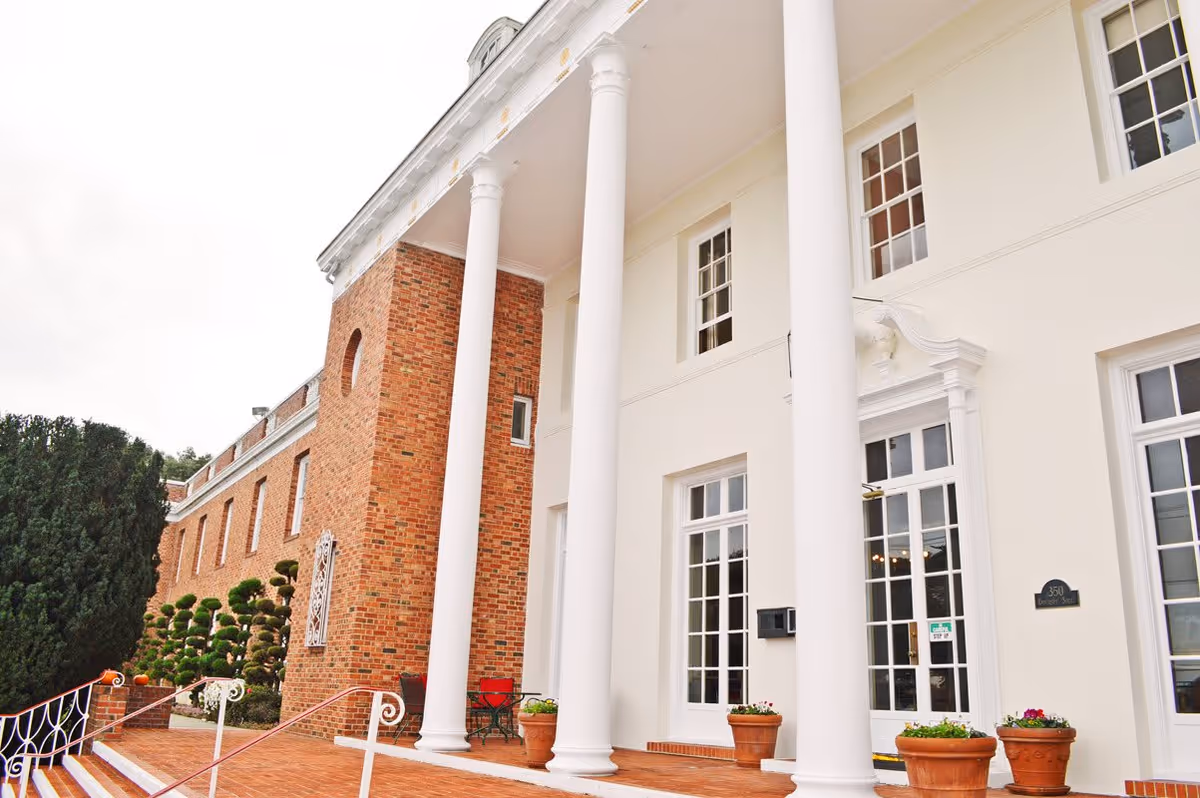 Exterior view of a senior living facility named Portola Gardens featuring a large white building with tall white columns, brick walls, multiple windows, a staircase with white railings, potted plants, and neatly trimmed bushes.