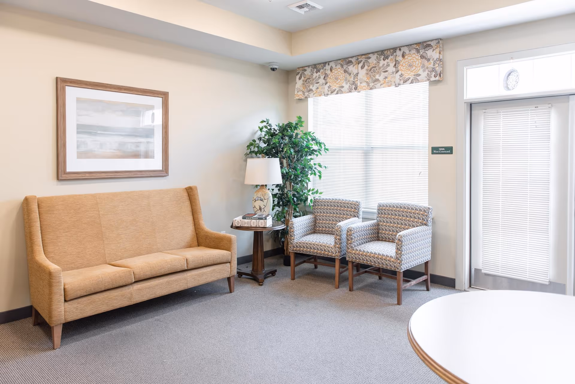 A bright and clean sitting area in a senior living facility with a beige three-seat sofa, two patterned armchairs, a small round wooden side table with a decorative lamp and books, a large potted plant, a framed abstract painting on the wall, and a window with floral valance and blinds next to a door with a window blind.