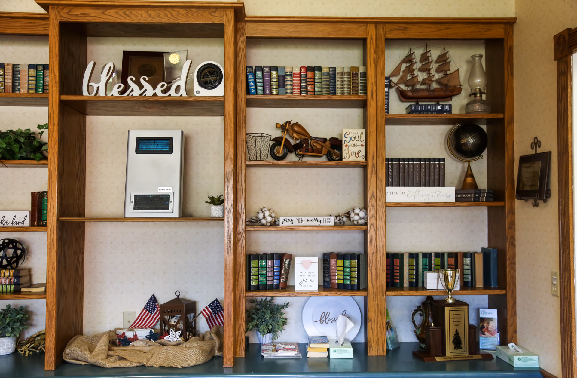 Wooden built-in shelves filled with decorative items including books, small American flags, a model ship, a globe, trophies, plants, and various inspirational signs such as 'blessed' and 'pray more worry less'.