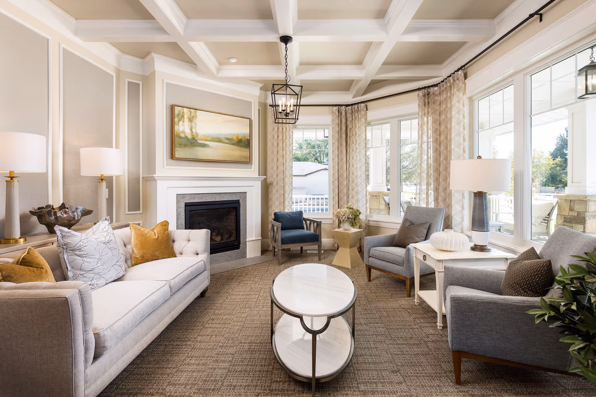 A bright and elegant living room with a beige sofa adorned with decorative pillows, two gray armchairs, a blue armchair, a white oval coffee table, and a white side table with a lamp and decorative pumpkin. The room features a fireplace with a landscape painting above it, large windows with sheer curtains, and a coffered ceiling with a black chandelier.