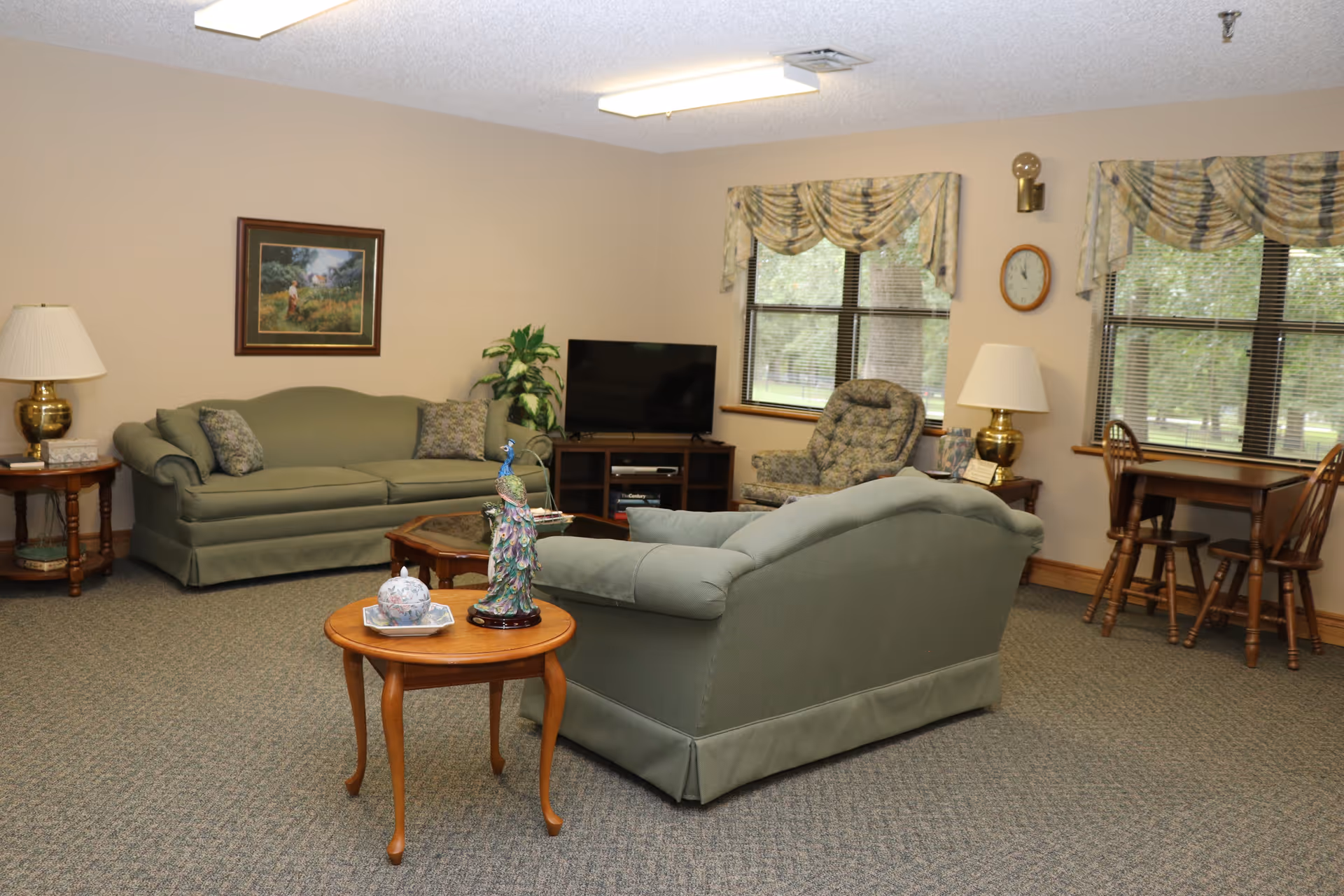 A cozy living room area with two green sofas, a patterned armchair, a wooden coffee table, and side tables with lamps. There is a flat-screen TV on a wooden stand, two windows with floral valances, a wall clock, and a small wooden dining table with chairs near the windows. The room has beige walls and carpeted flooring.
