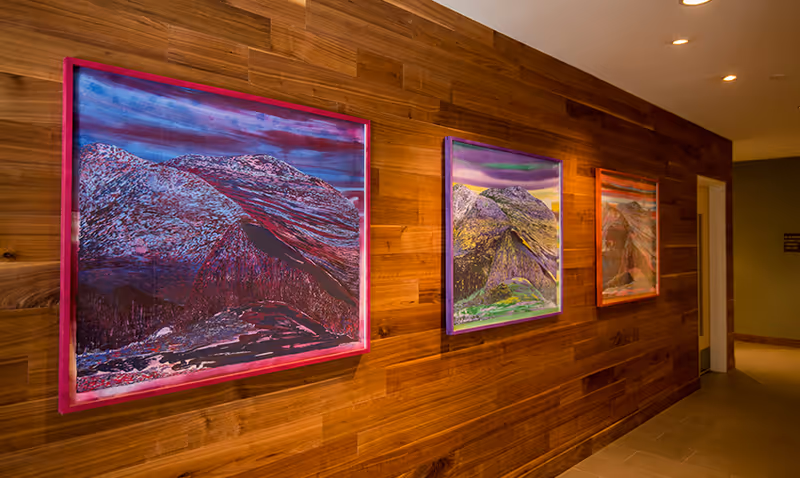 A hallway with wooden paneled walls displaying three colorful framed paintings of mountainous landscapes. The floor is tiled and the ceiling has recessed lighting.