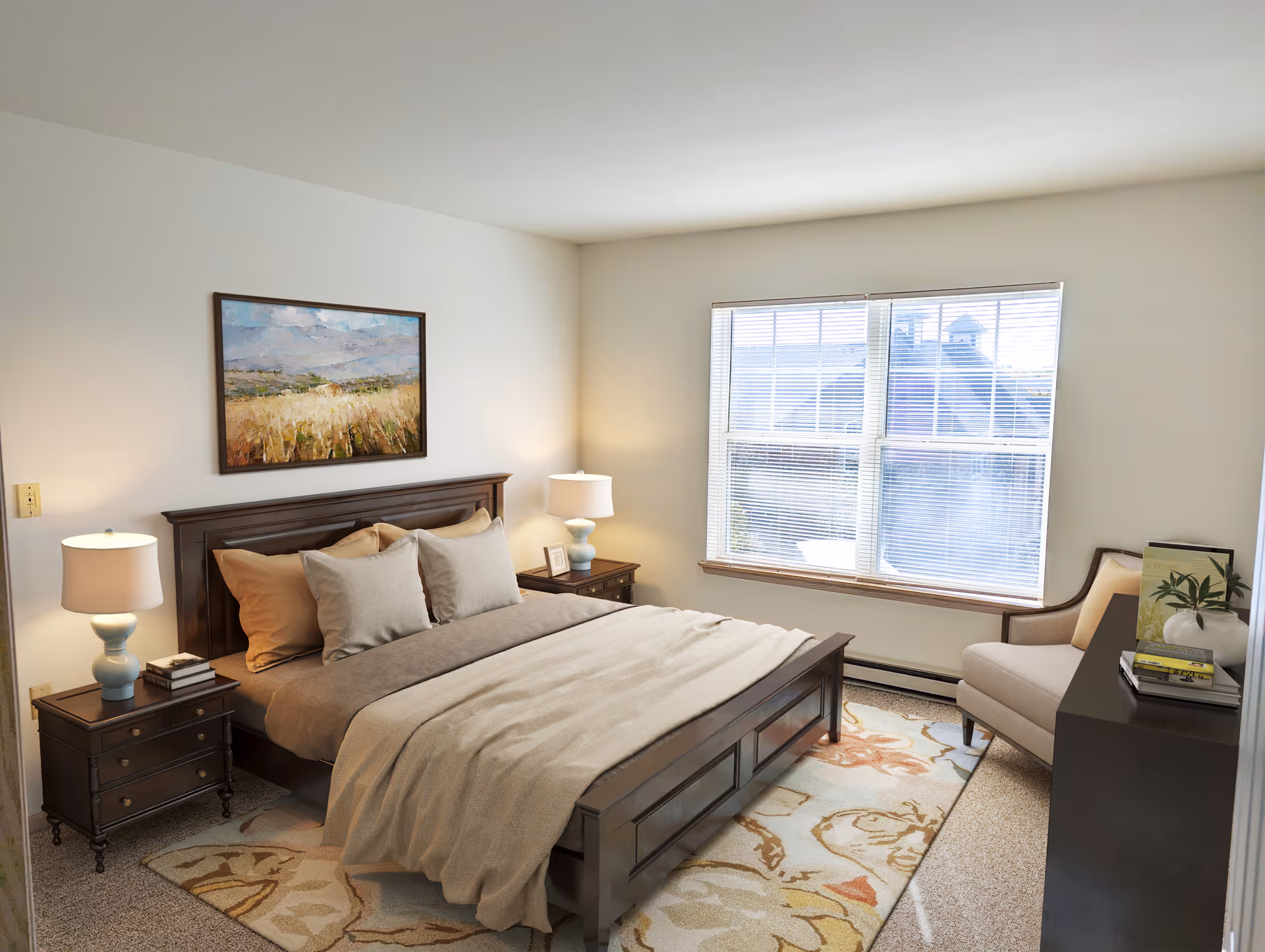 A cozy bedroom with a large wooden bed featuring beige and brown bedding, two matching nightstands with lamps, a framed landscape painting above the bed, a window with blinds letting in natural light, a cushioned armchair, and a dark dresser with books and a plant on top.