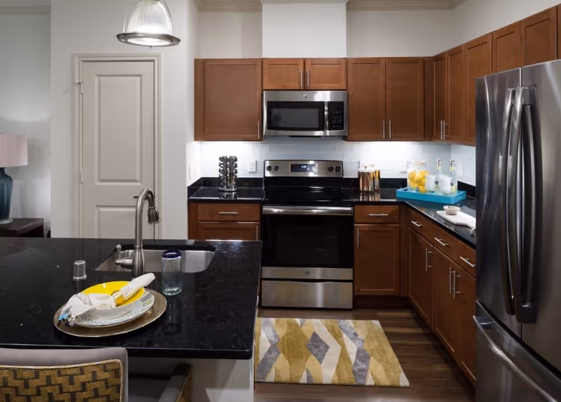 Modern kitchen with dark granite countertops, wooden cabinets, stainless steel appliances including a refrigerator, oven, and microwave. A kitchen island with a sink and a plate with utensils is in the foreground. A colorful patterned rug is on the wooden floor in front of the oven.