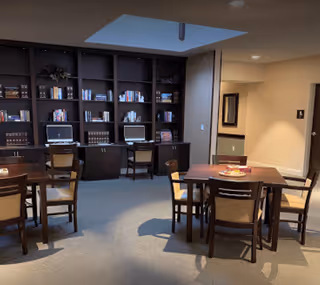 A quiet room with several wooden tables and chairs arranged for seating. Along one wall is a large built-in bookshelf filled with books and decorative items, with two computer workstations integrated into the shelving. The room is softly lit with recessed ceiling lights and has a skylight providing natural light.