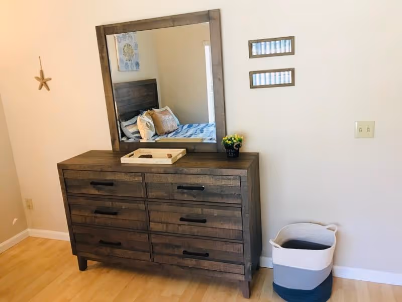 A wooden dresser with six drawers and a large matching mirror on top. On the dresser, there is a small tray and a small potted plant. To the right of the dresser is a white, gray, and blue woven basket. The reflection in the mirror shows a bed with pillows and a headboard. The room has light-colored walls and wooden flooring.