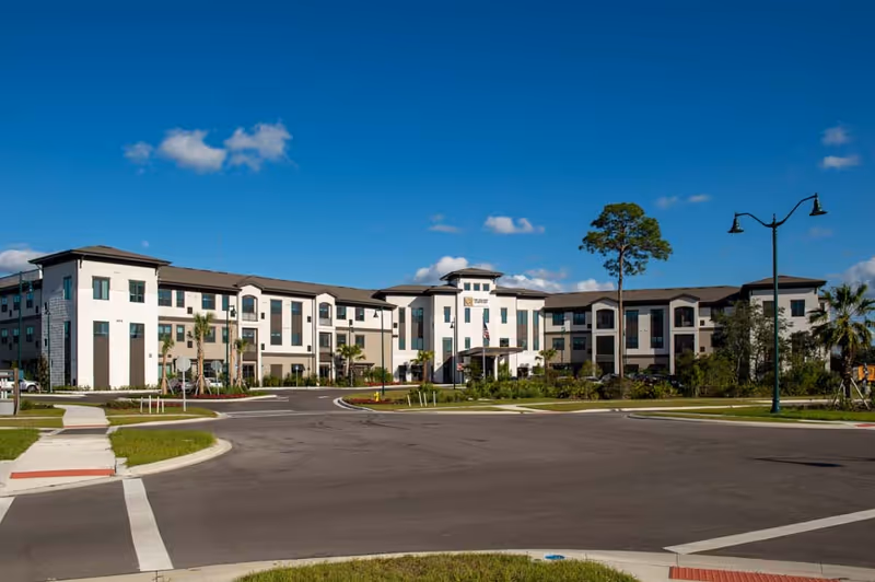 Front view of The Gallery at North Port, a three-story senior living building with landscaped grounds under a clear blue sky.