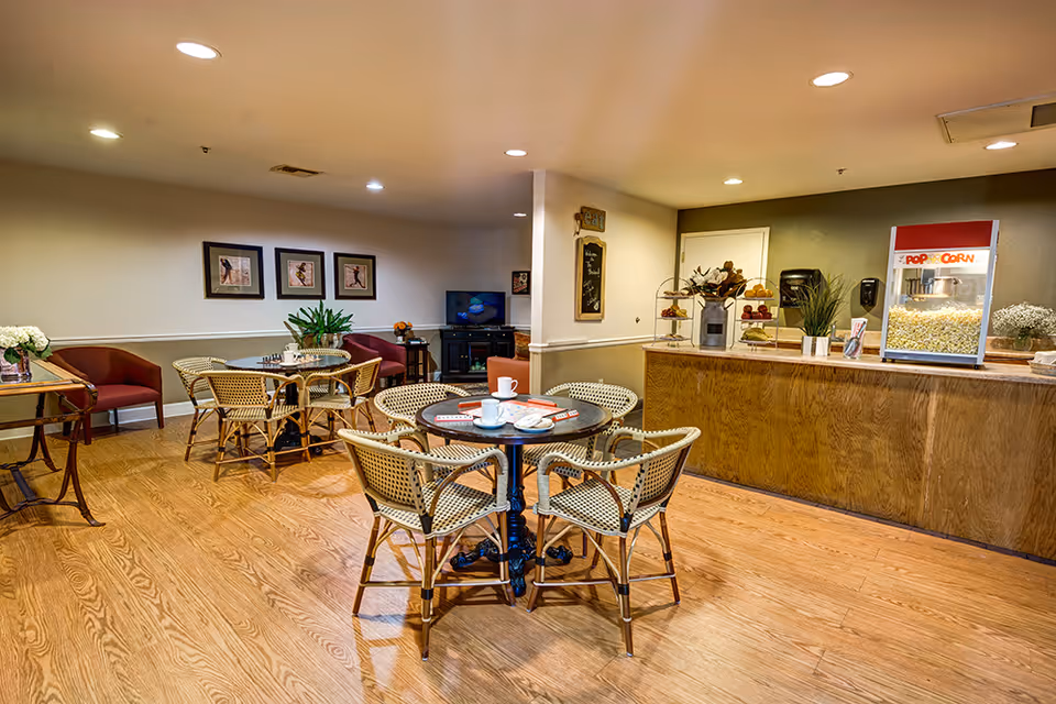 A cozy common area with wooden flooring featuring round tables surrounded by wicker chairs. A popcorn machine sits on a wooden counter to the right, and a TV is visible in the background. The walls are decorated with framed pictures and plants, creating a welcoming atmosphere.