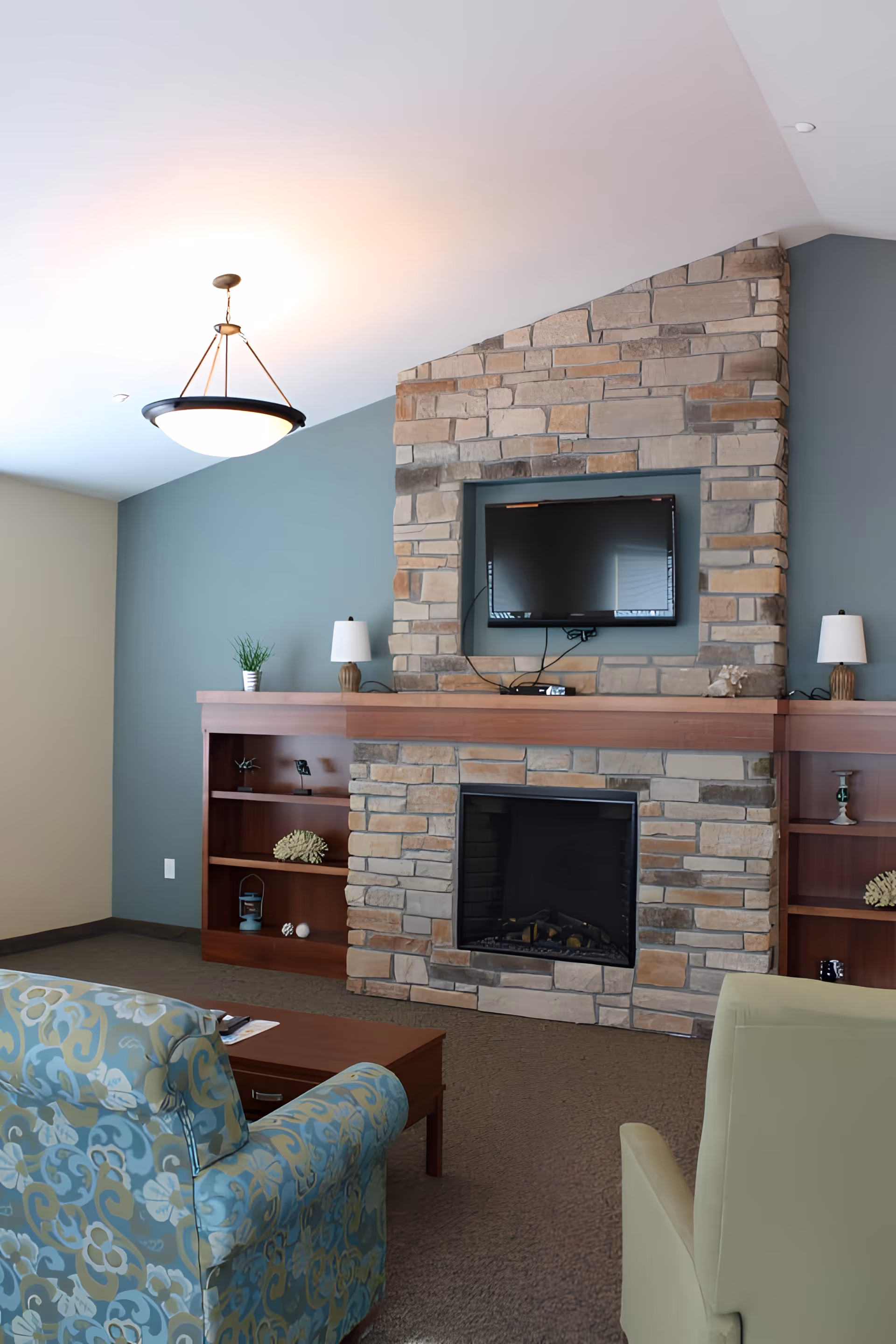 A cozy living room area featuring a stone fireplace with a mounted flat-screen TV above it. On either side of the fireplace are wooden built-in shelves decorated with small plants, lamps, and decorative items. In front of the fireplace are two armchairs, one with a blue and green floral pattern and the other a solid light green color, along with a wooden coffee table. The walls are painted in a soft blue and beige color, and a ceiling light fixture hangs above.