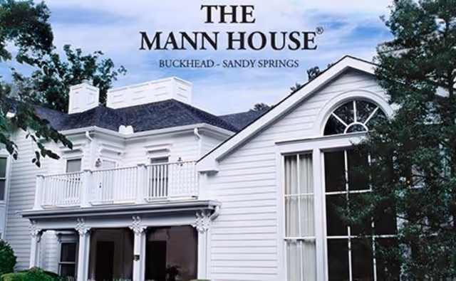 Exterior view of a white two-story house with a balcony and large windows, surrounded by trees and greenery under a blue sky. The text 'THE MANN HOUSE BUCKHEAD - SANDY SPRINGS' is displayed above the house.