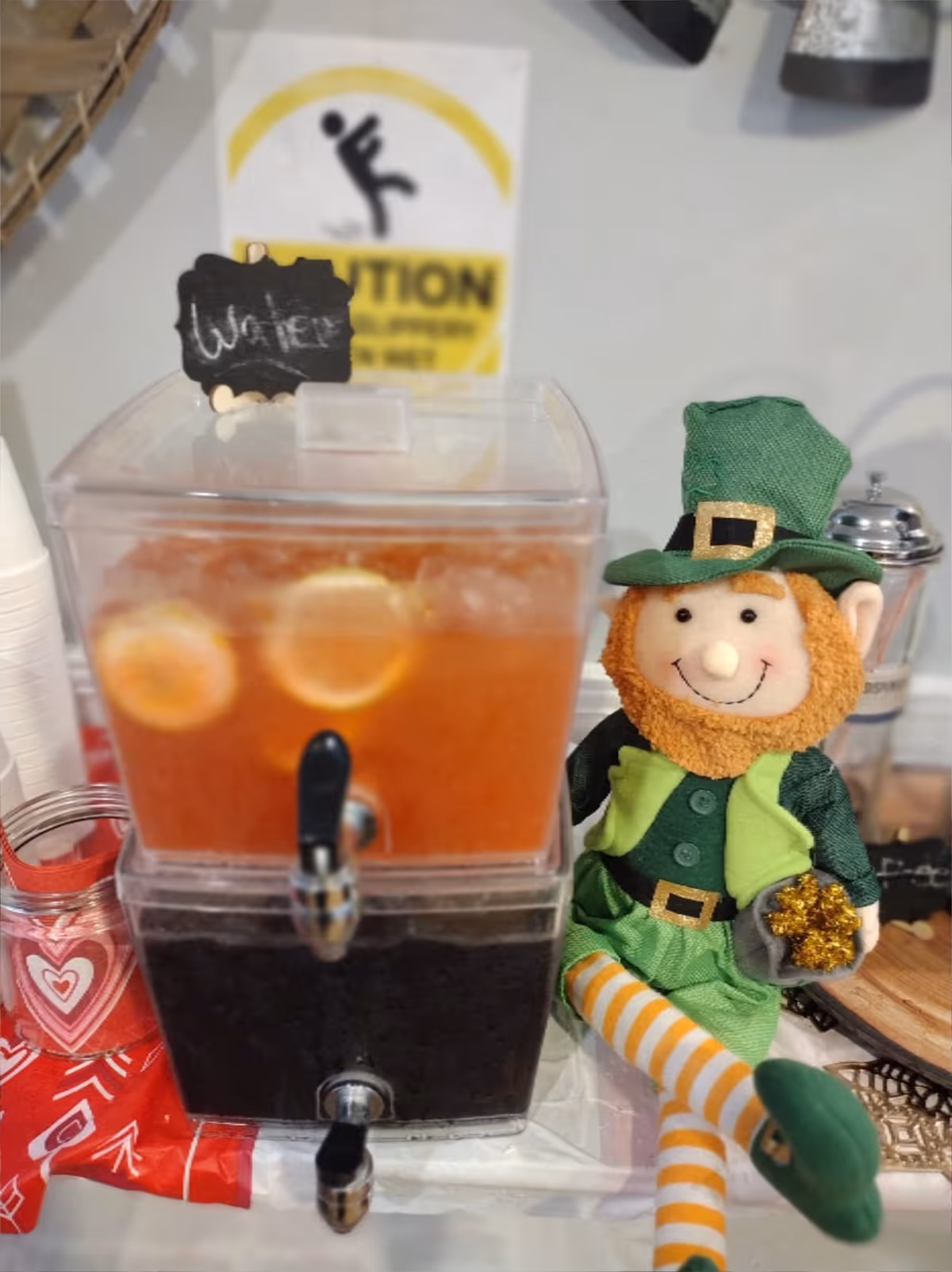 Two-tier beverage dispenser with an orange-slice drink and a dark beverage next to a smiling leprechaun doll on a table.