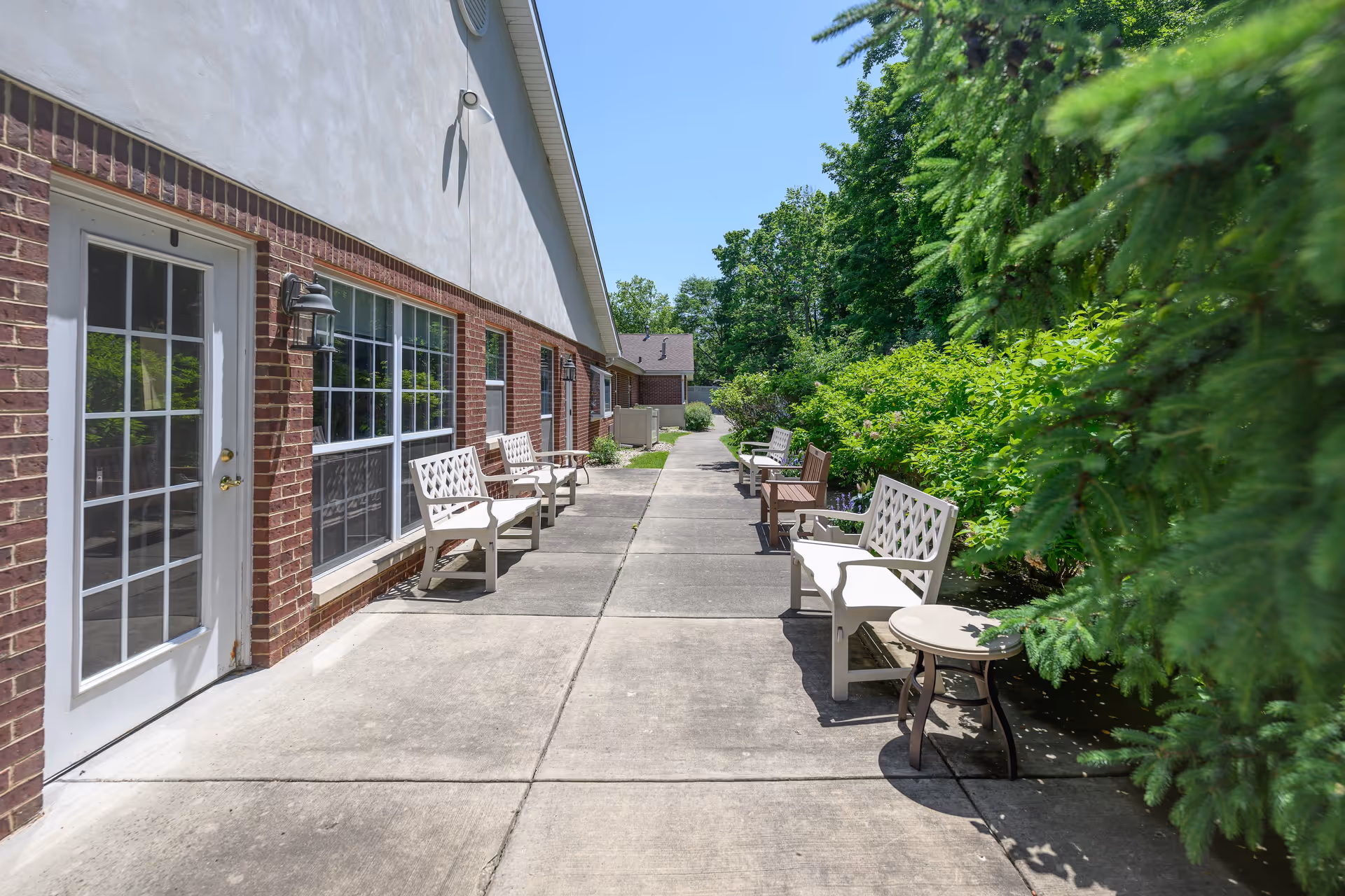 Outdoor walkway alongside a brick building with multiple white benches and a small round table. The walkway is bordered by green bushes and trees under a clear blue sky.