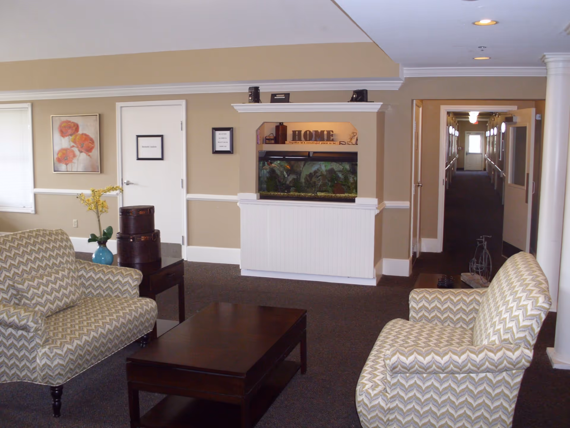 A cozy sitting area in a senior living facility with two patterned armchairs facing a dark wooden coffee table. Behind the seating area is a beige wall with a built-in fish tank and a decorative sign that reads 'HOME'. To the left is a door labeled 'Beauty Salon' and a framed floral painting. A hallway extends to the right with several doors and handrails along the walls.