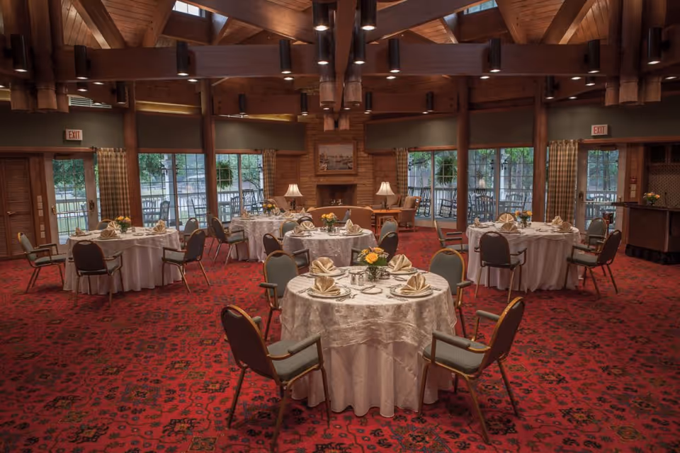 A spacious dining room with round tables covered in white tablecloths, each set with folded napkins, plates, and silverware. The room features a red patterned carpet, wooden beams on the ceiling, large windows with views of an outdoor patio, and a fireplace with lamps on either side.