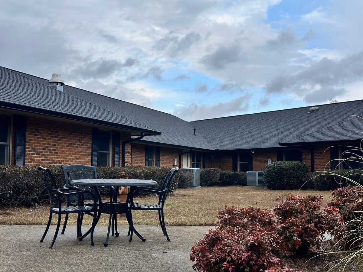 Metal patio table and chairs on a concrete courtyard with shrubs and a one-story brick building in the background under a cloudy sky.