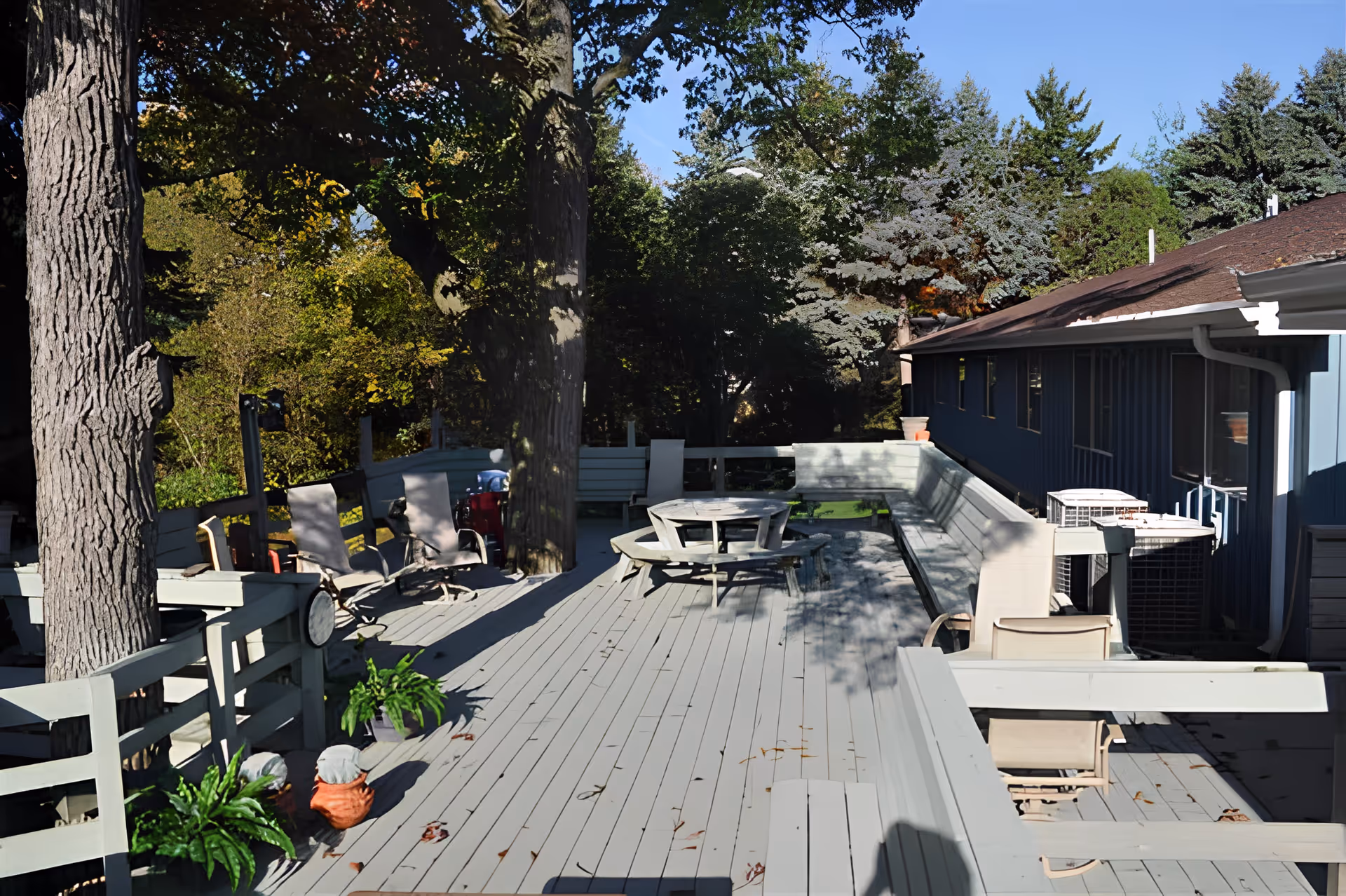 Outdoor wooden deck area with several chairs, a round picnic table with attached benches, potted plants, and large trees providing shade. The deck is adjacent to a blue building with multiple windows and air conditioning units. The surrounding area has dense green trees under a clear blue sky.