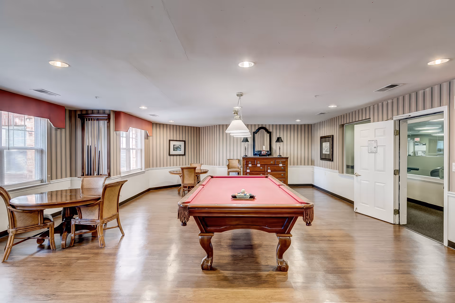 A spacious recreational room with a red felt pool table in the center, surrounded by wooden chairs and round tables. The room has striped wallpaper, wooden flooring, and several windows with red valances letting in natural light. A wooden dresser with a mirror and lamp is against the far wall, and an open door leads to another room.