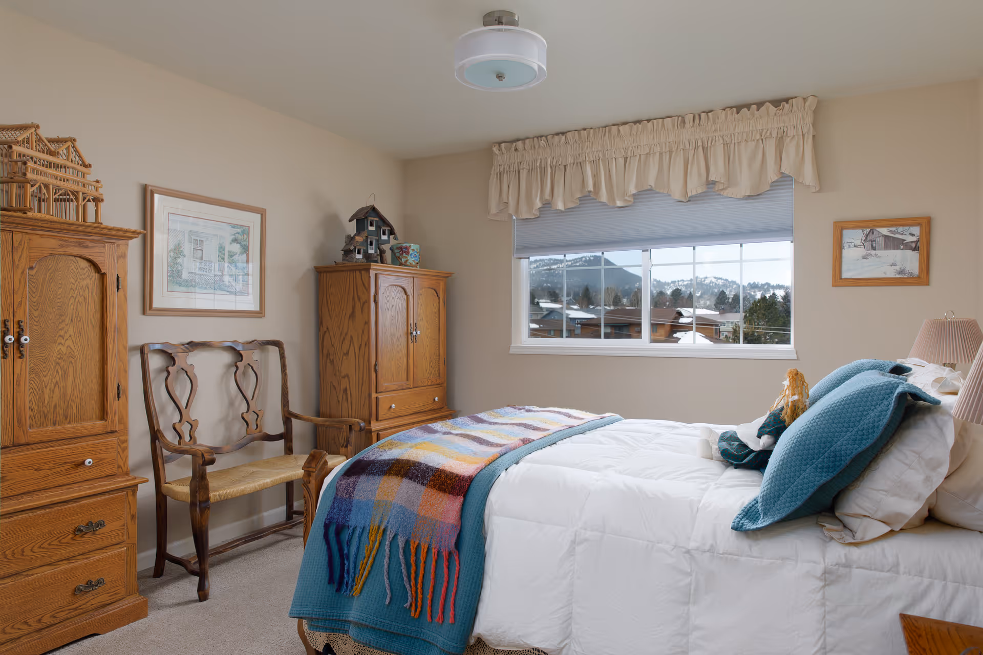 A cozy bedroom with a neatly made bed featuring white bedding and teal pillows, a colorful plaid throw blanket, wooden furniture including a cabinet and a bench, a window with a view of snow-covered rooftops and trees, and framed artwork on the walls.