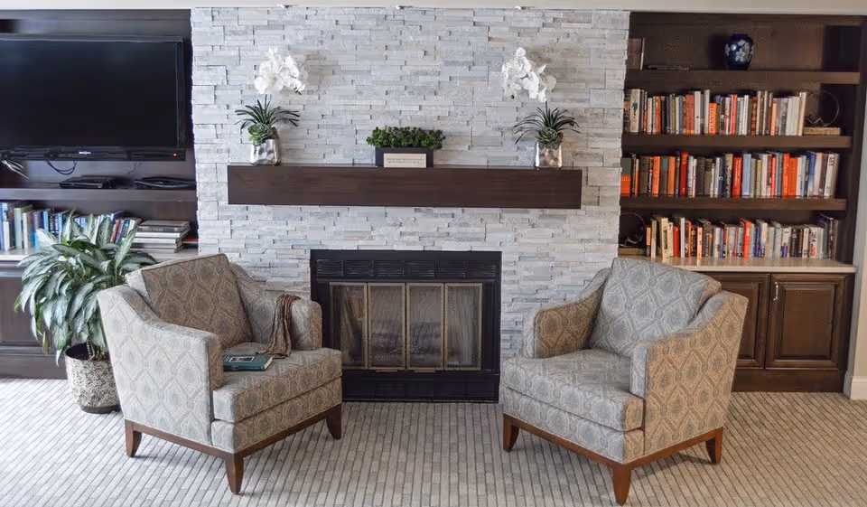A cozy living room area with two patterned armchairs facing each other in front of a stone fireplace. Above the fireplace is a wooden mantel with two white orchid plants and a small green plant. On either side of the fireplace are built-in dark wood shelves filled with books and decorative items. A flat-screen TV is mounted on the left side above the shelves, and a potted plant is placed on the floor next to the left armchair.