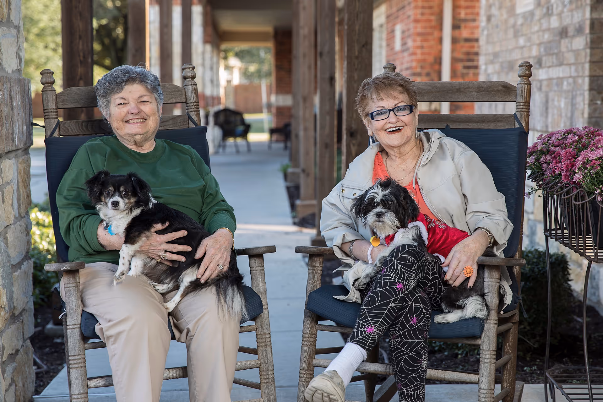 Two elderly women sitting on wooden rocking chairs outside a brick building, each holding a small dog. They are smiling and appear to be enjoying a pleasant day outdoors.