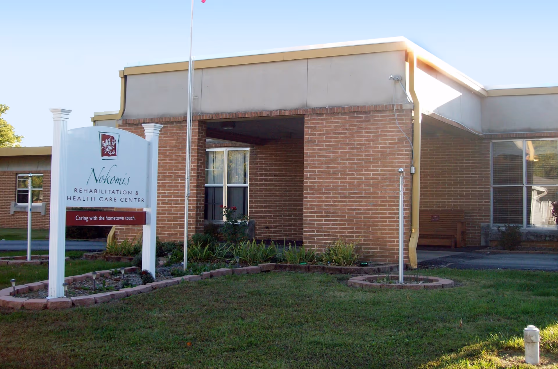 Exterior view of Nokomis Rehabilitation & Health Care Center showing a brick building with windows, a covered entrance, a flagpole, and a white sign with the facility's name and slogan 'Caring with the hometown touch.' There is a grassy area and some landscaping in front of the building.