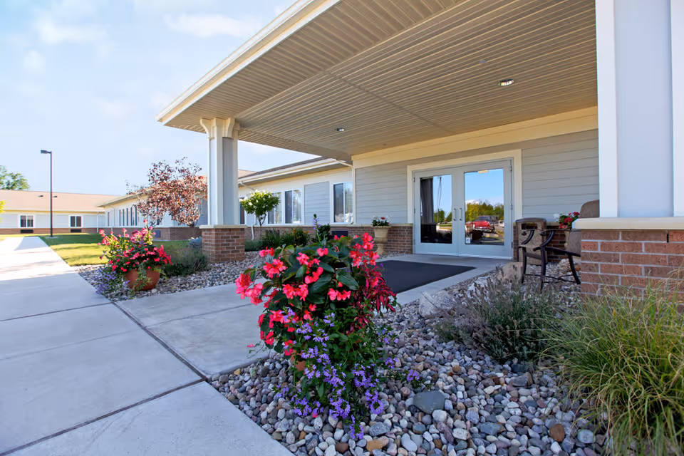 Entrance of a senior living facility with a covered porch, double glass doors, and a landscaped area featuring colorful flowers and rocks. There is a sidewalk leading to the entrance and a chair placed near the door.