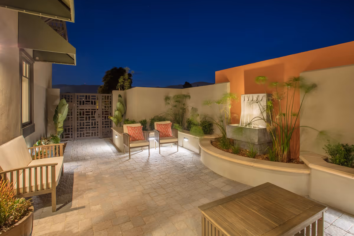 Well-lit outdoor courtyard at night with patio chairs, planters, and a decorative wall fountain.