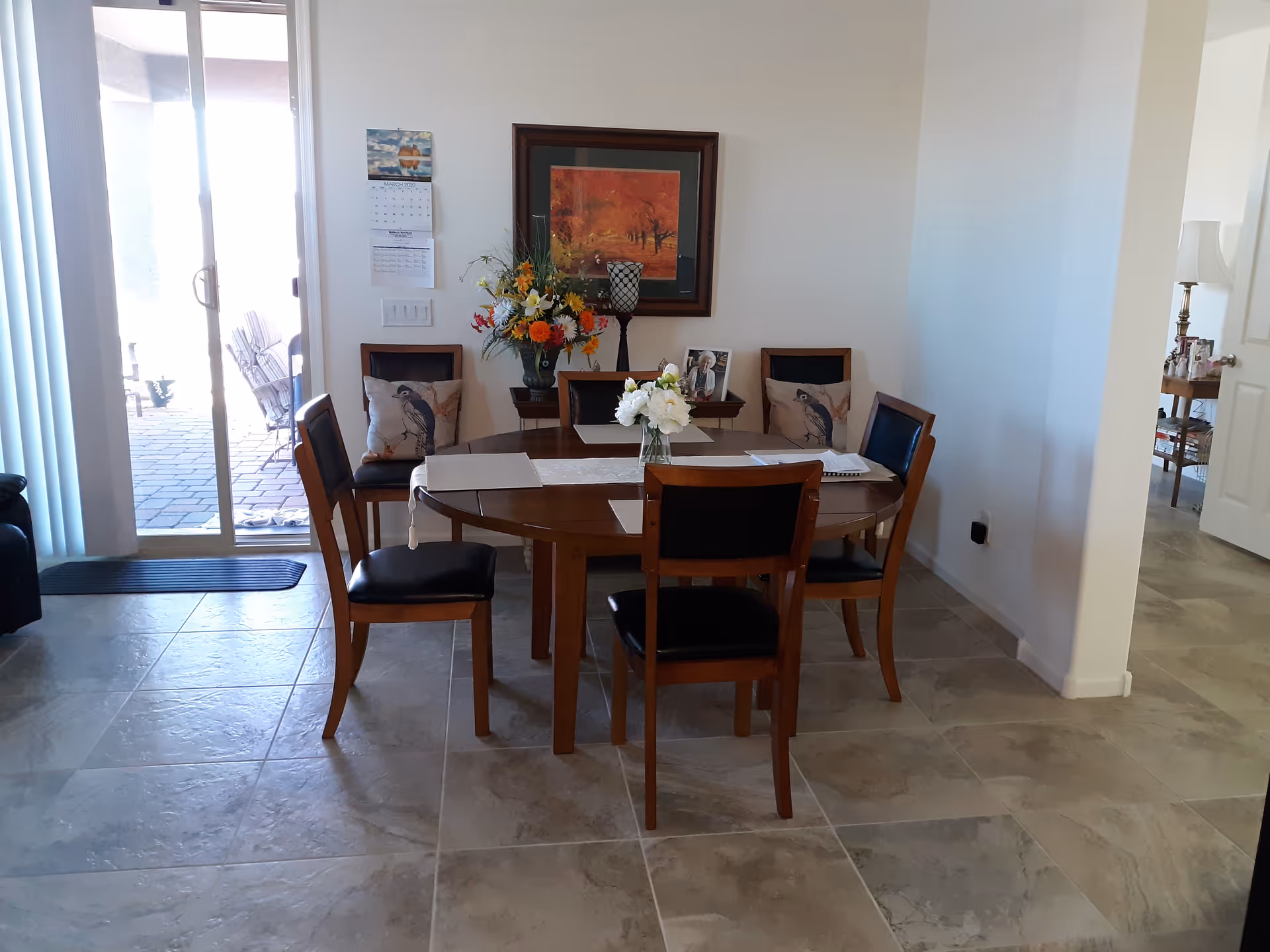 A dining area with a wooden oval table surrounded by six chairs with black cushions. The table has a white runner and a vase with white flowers in the center. Behind the table is a small side table with a colorful flower arrangement, a framed photo, and a decorative candle holder. A framed painting with autumn colors hangs on the wall above the side table. To the left, there is a sliding glass door with vertical blinds leading to an outdoor patio with chairs. The floor is tiled with large beige tiles.