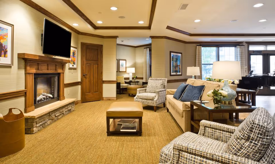 A cozy living room area in a senior living facility with a beige sofa, patterned armchairs, a brown ottoman, and a wooden side table with a lamp and flowers. There is a fireplace with a mounted flat-screen TV above it, framed artwork on the walls, and large windows letting in natural light. The room has warm lighting and a carpeted floor.