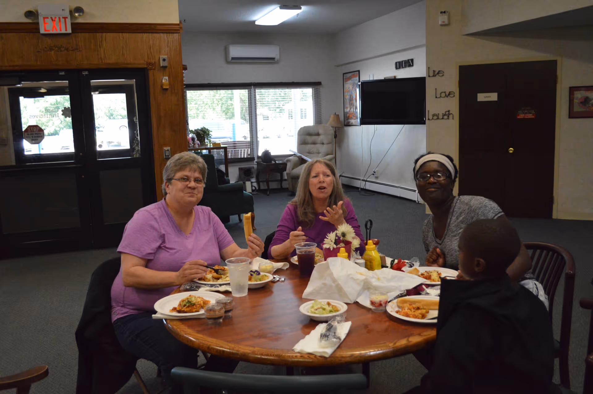 Four people sitting around a round wooden table inside a common area, eating a meal together. The room has large windows, a TV mounted on the wall, and a door with an exit sign above it. The walls have decorations including a 'Live Love Laugh' sign.