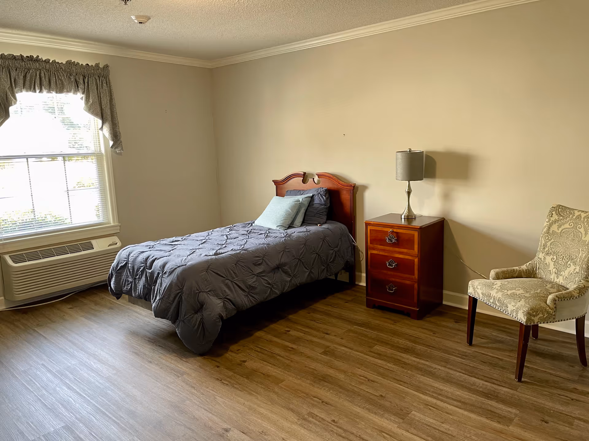 A simple bedroom with a single bed covered in a gray comforter and two pillows. Next to the bed is a wooden nightstand with three drawers and a lamp on top. To the right of the nightstand is an upholstered armchair with a patterned fabric. A window with blinds and a valance is on the left wall, and there is a wall-mounted air conditioning unit below the window. The floor is wood, and the walls are painted a light beige color.