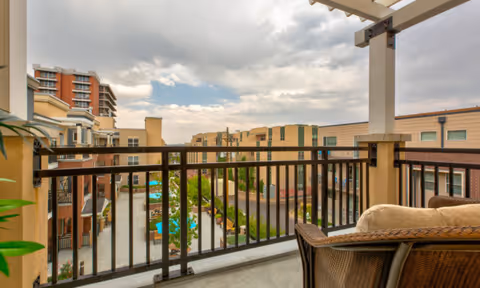 View from a balcony overlooking a courtyard with a swimming pool, surrounded by multi-story residential buildings under a cloudy sky. The balcony has a black metal railing and a wicker chair with a cushion.