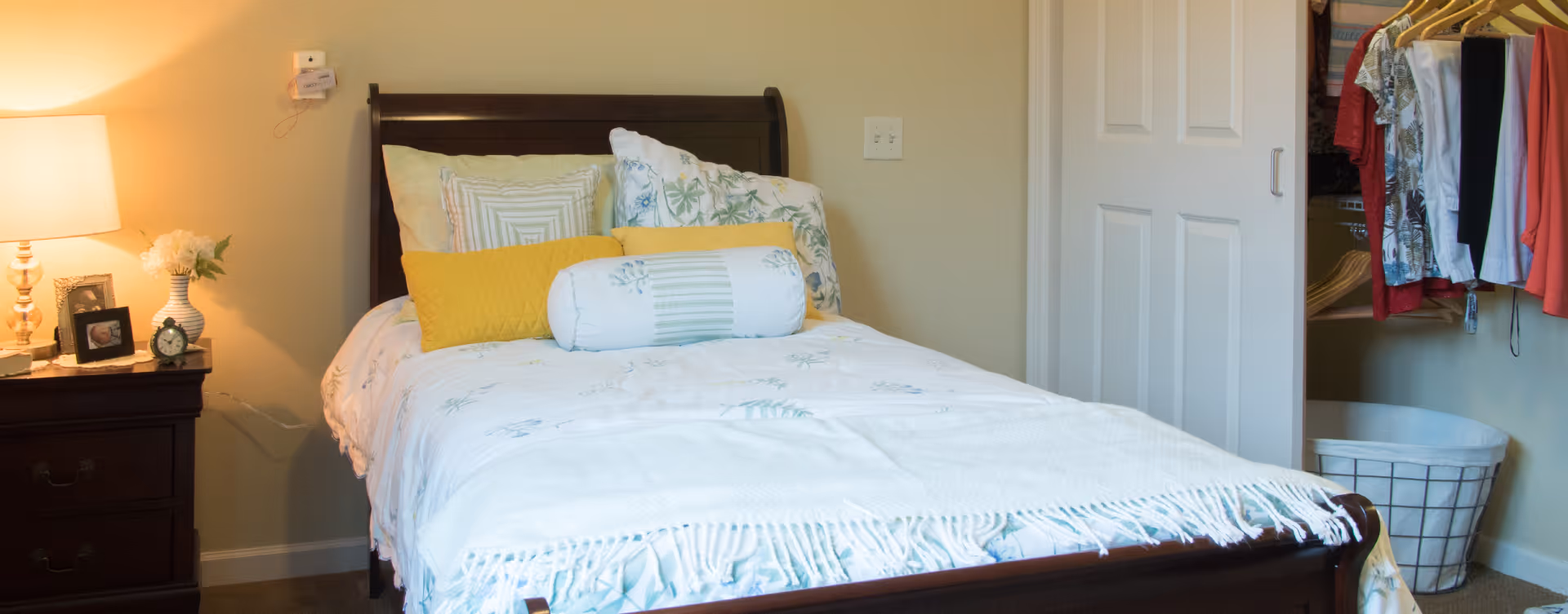 A cozy bedroom featuring a wooden bed with white and floral bedding, accented by yellow and white pillows. To the left of the bed is a dark wooden nightstand with a lamp, a small clock, a framed photo, and a vase with flowers. On the right side, there is a closet with sliding white doors, partially open to reveal hanging clothes and a laundry basket.