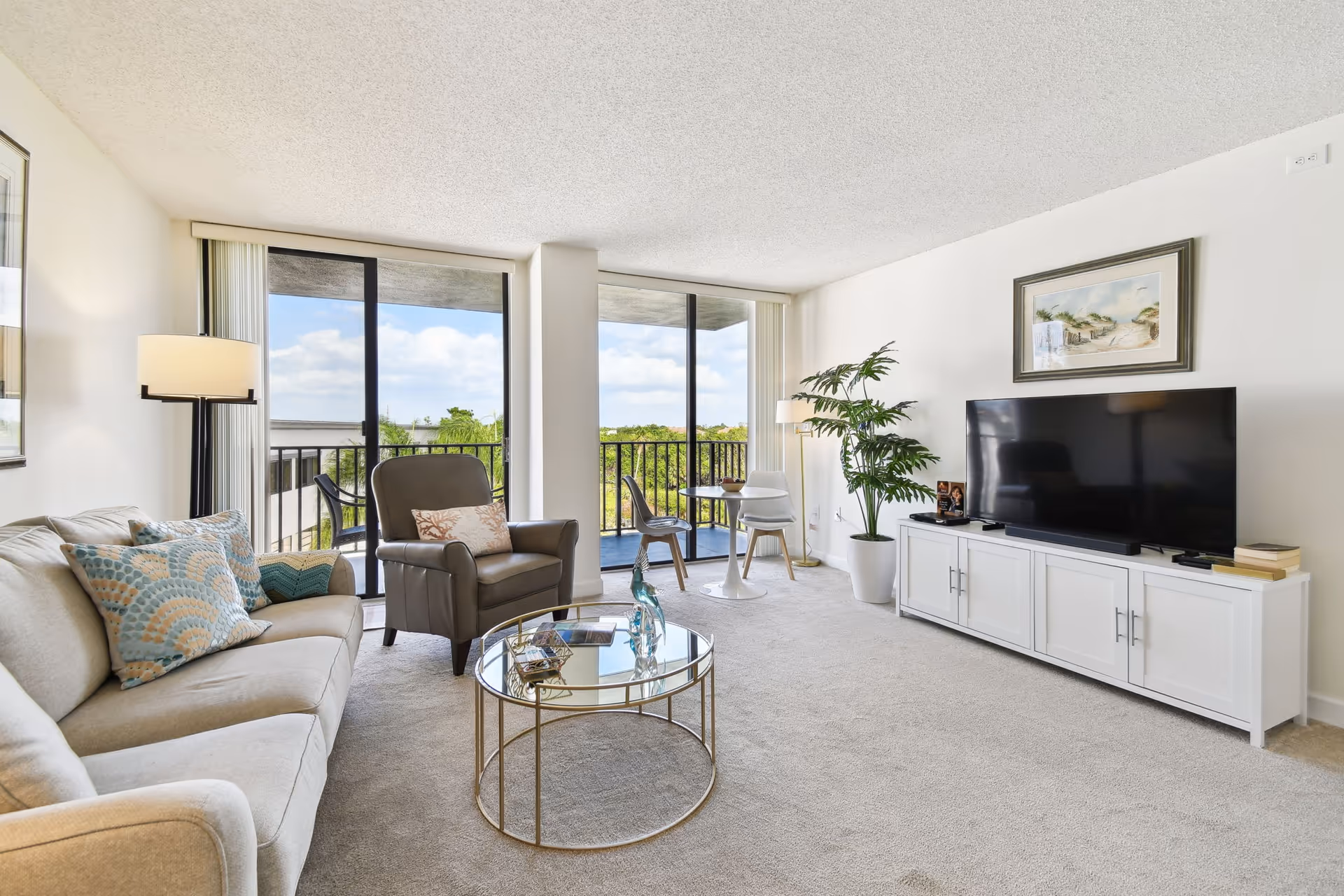 A bright living room with a beige sofa adorned with patterned cushions, a dark armchair with a decorative pillow, and a round glass coffee table with decorative items. The room features large sliding glass doors leading to a balcony with outdoor seating and a view of greenery. A white TV cabinet holds a large flat-screen television, with a potted plant and framed artwork on the wall above it. The room is carpeted and has a floor lamp near the sofa.