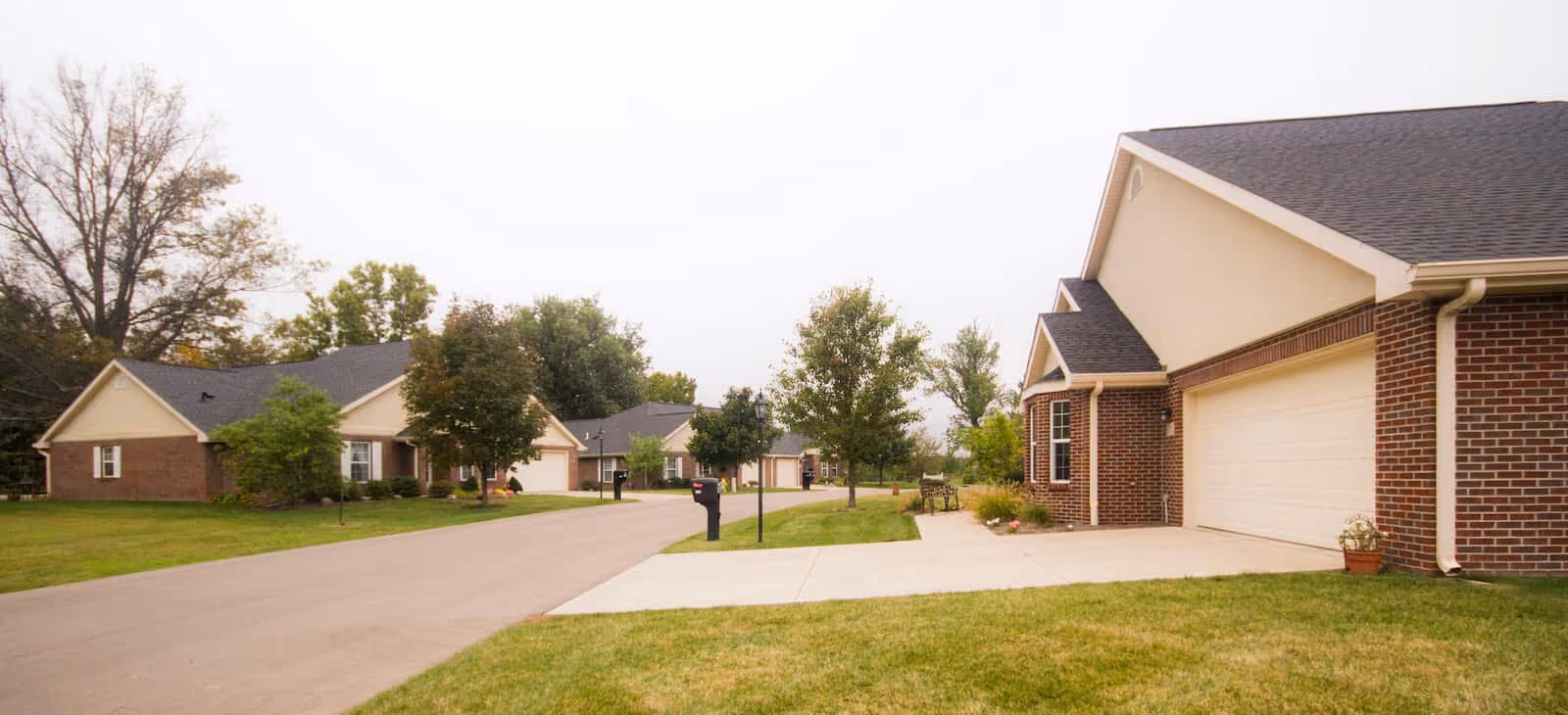 Quiet suburban street lined with single-story brick houses, garages, driveways, and lawns under an overcast sky.