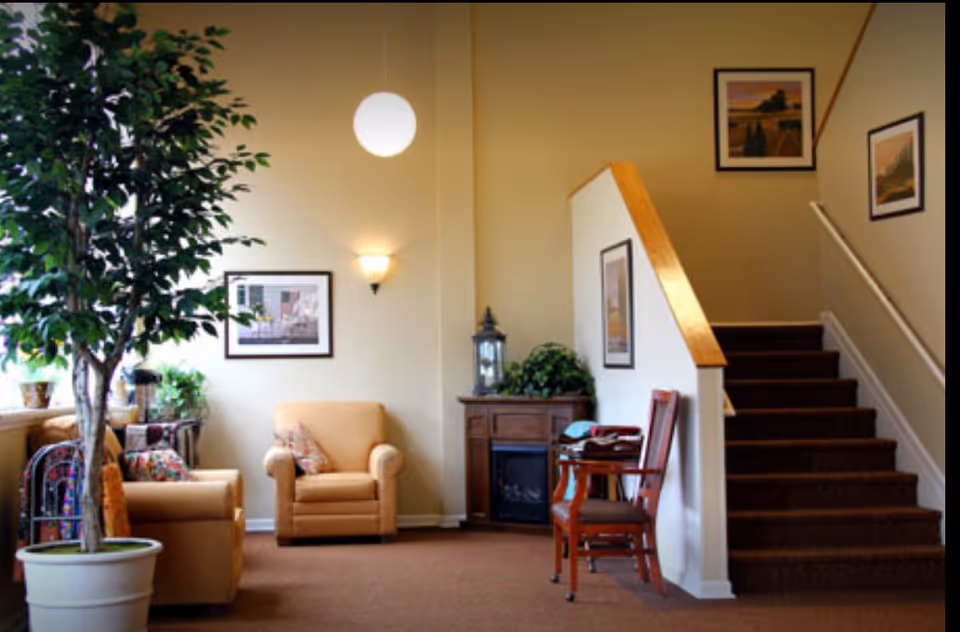 A cozy senior living facility common area with beige armchairs, a potted tree, framed artwork on the walls, a small wooden cabinet with a decorative lantern and plant, and a carpeted staircase with wooden handrails.