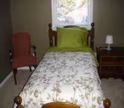 A small bedroom with a single wooden bed made with a green pillow and floral comforter, a nightstand and lamp, and an upholstered chair by a window.