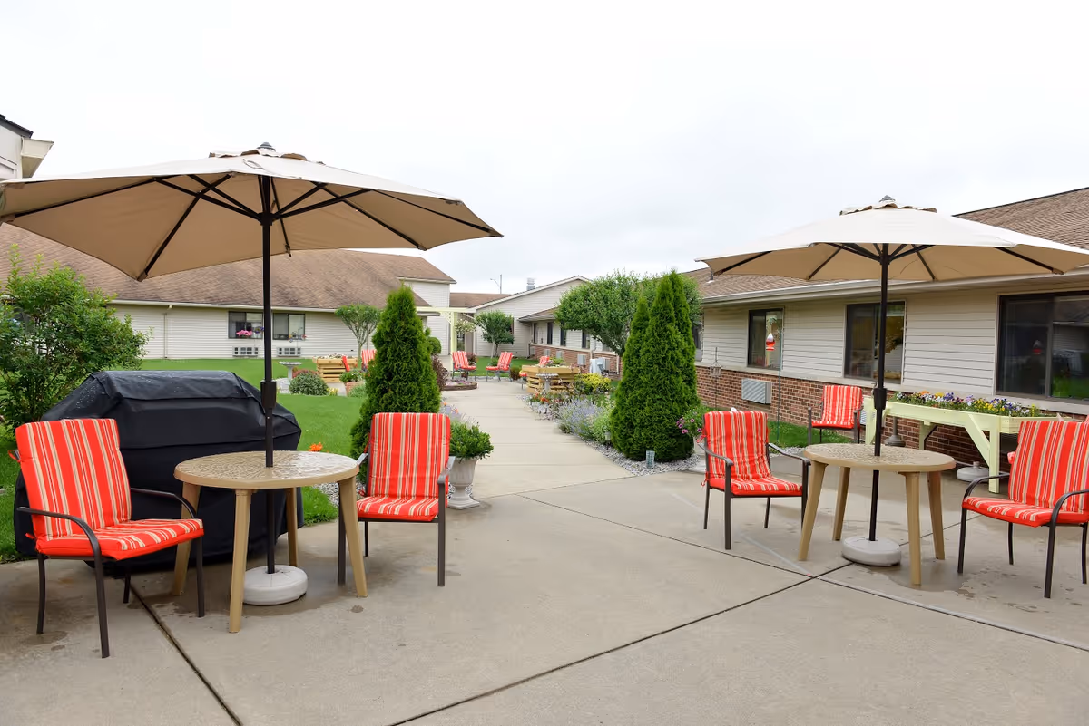 Outdoor patio area at The Laurels of Defiance with two round tables each shaded by large beige umbrellas. Each table is surrounded by red and white striped cushioned chairs. There is a black covered grill on the left side and various green shrubs and flowers along the walkway between single-story buildings.