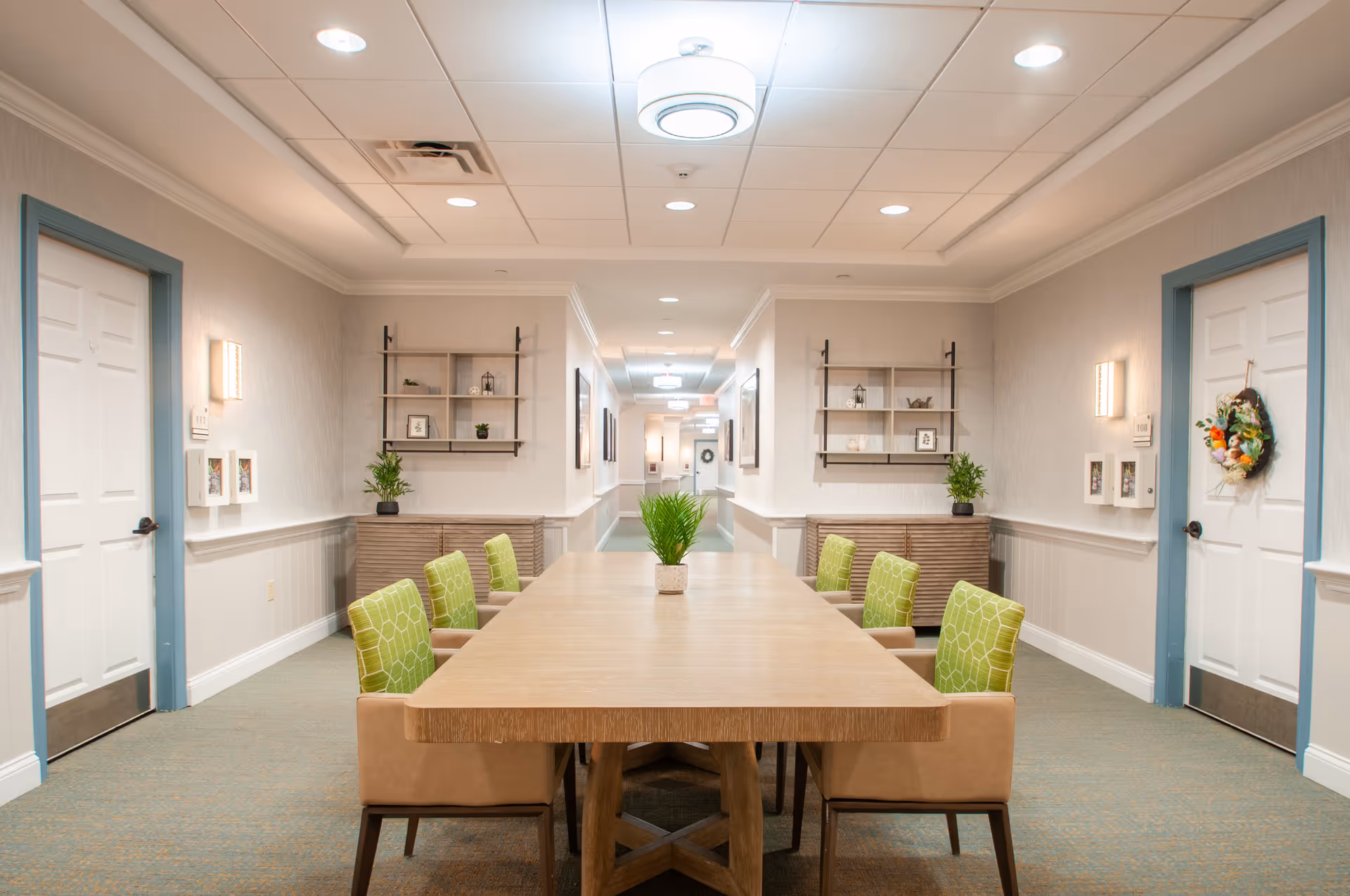 A long wooden table with eight chairs featuring green patterned cushions is centered in a well-lit hallway with light-colored walls and ceiling. Two white doors with blue trim are visible on either side of the hallway, each decorated with small wall-mounted shelves and plants. The hallway extends into the distance with additional lighting fixtures on the ceiling.