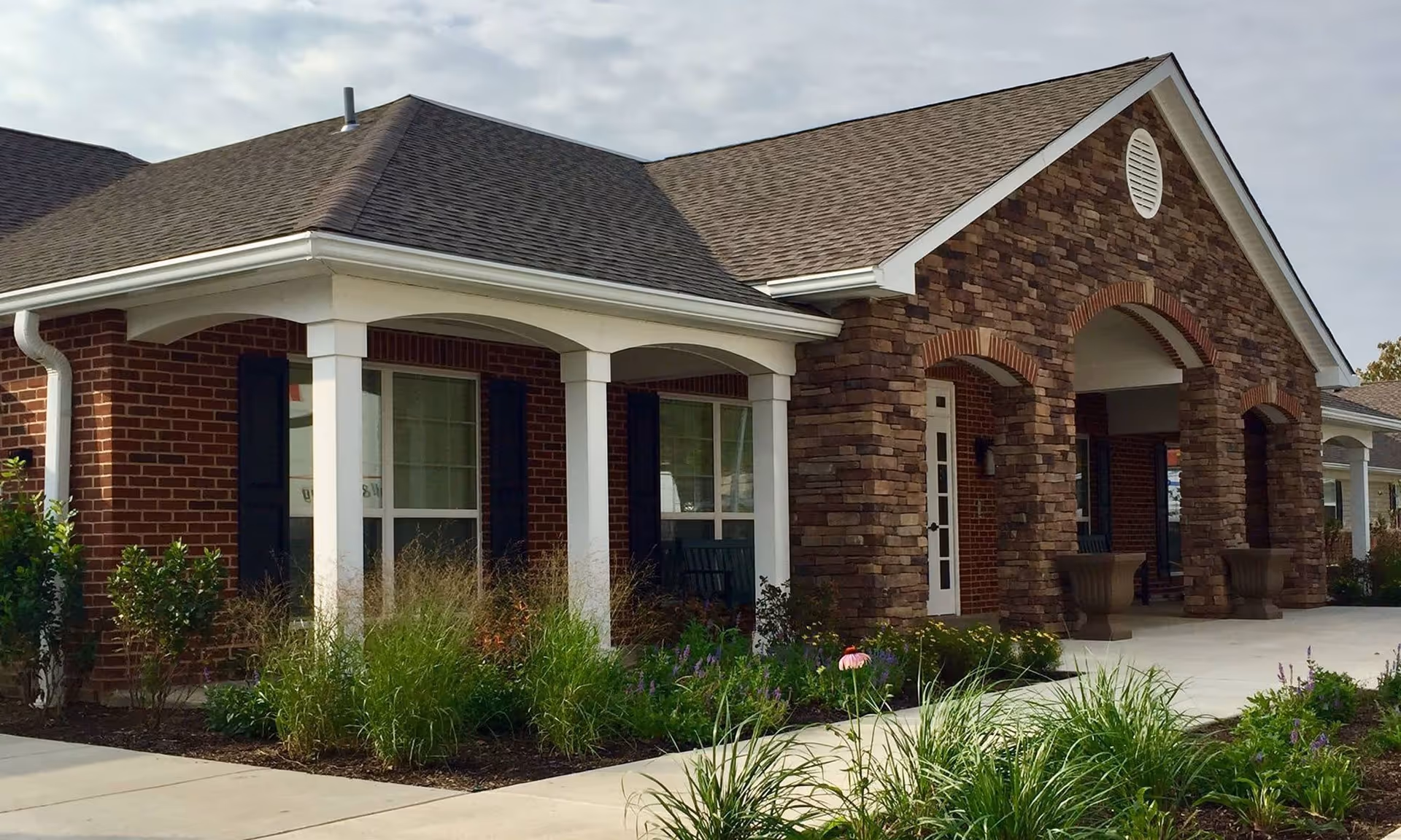 Exterior view of a senior living facility building with a combination of brick and stone facade, white columns, and a covered entrance. There are plants and shrubs along the walkway leading to the entrance.