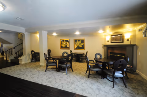 Interior view of a common area in a senior living facility with round tables and chairs arranged on a carpeted floor. The room features a fireplace with a framed picture above it, two framed leaf artworks on the wall, and a staircase visible in the background.