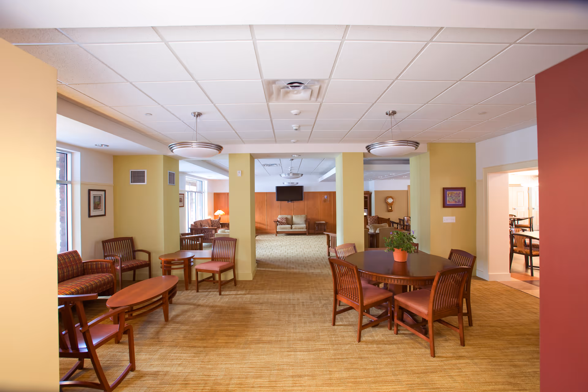 A spacious and well-lit common area in a retirement residence featuring multiple seating arrangements including wooden chairs with cushions, small tables, and a round table with a potted plant. The room has beige walls with some yellow and red accents, carpeted floors, and ceiling lights. In the background, there is a wall-mounted television and additional seating.