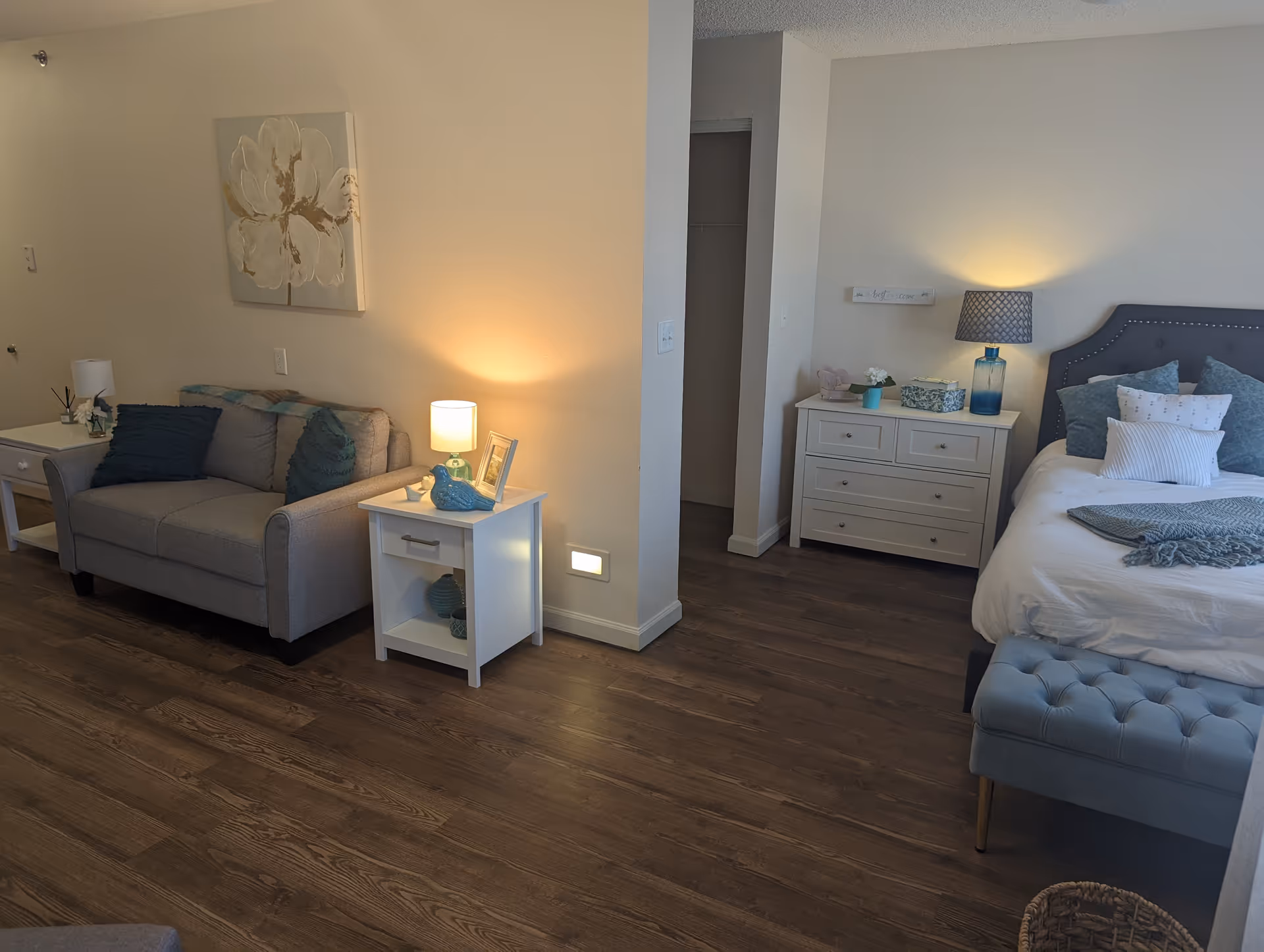 Studio-style interior with a bed, nightstands and dresser on the right and a small sofa with side tables and lamps on the left.