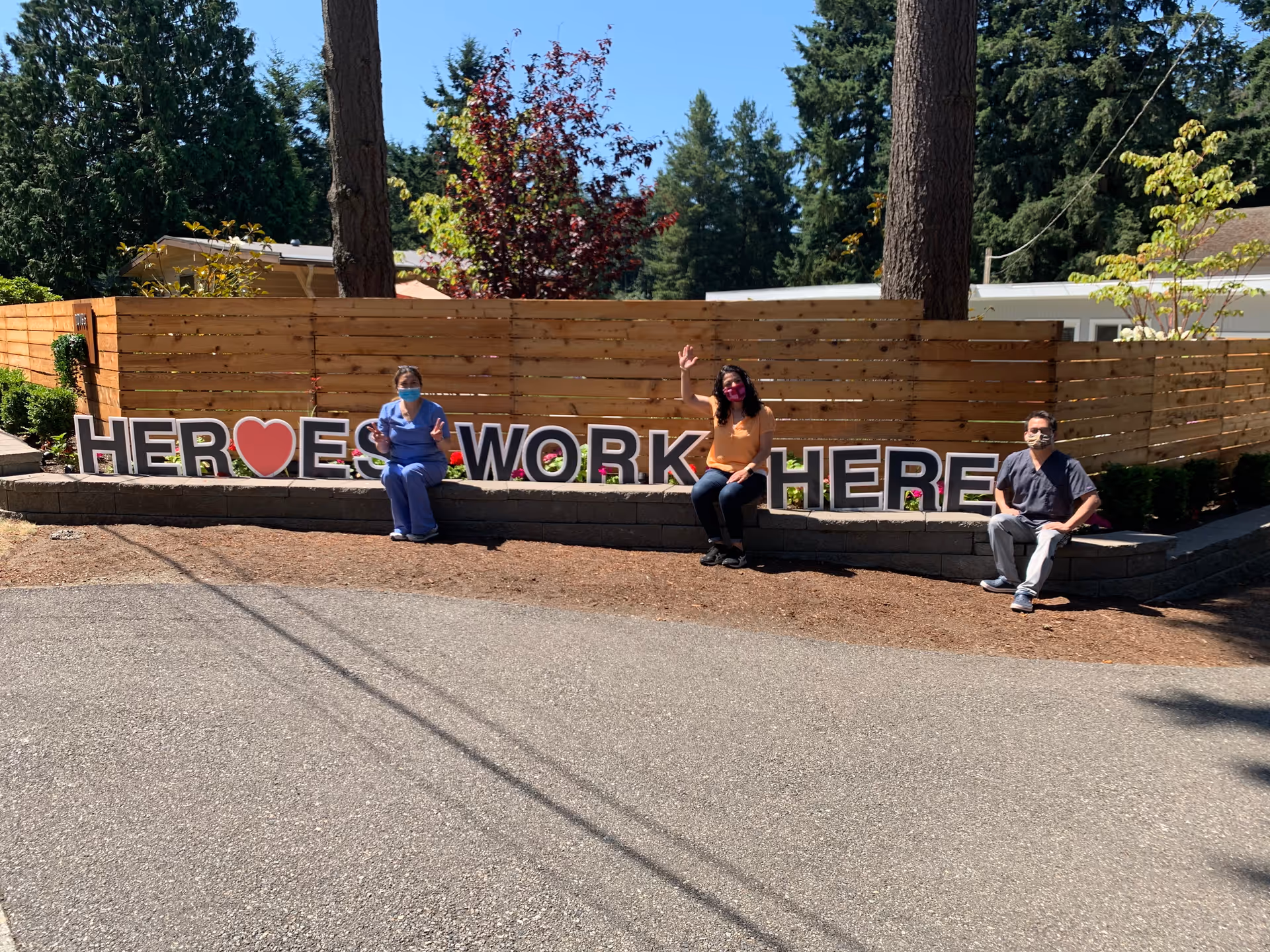 Three people wearing masks sitting spaced apart on a low stone wall in front of a wooden fence with trees in the background. Large letters on the wall spell out 'HEROES WORK HERE' with a red heart symbol replacing the letter O in HEROES. The people are waving and appear to be healthcare or essential workers.