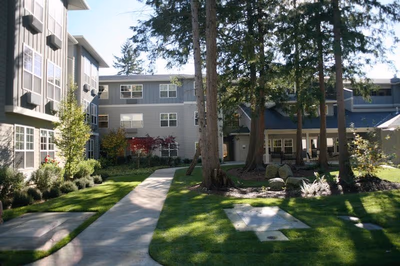 Outdoor view of a senior living facility courtyard with a paved walkway, green grass, trees, and multi-story residential buildings in the background under a clear sky.